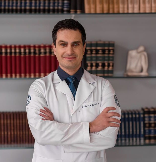 Dr. Marco Abud de jaleco no consultório, em frente a uma estante de livros